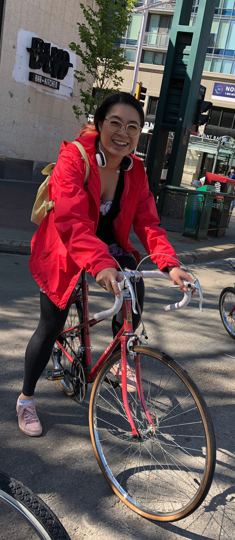 Photo shows a woman on a bike in the middle of an event in a downtown street. Serena has black hair, a red windbreaker, and black leggings on. The bike is reddish pink with white handles. She is looking at the camera and smiling. The event is in the afternoon, with blue skies and clouds showing a comfortable summer day. Downtown buildings are in the background.