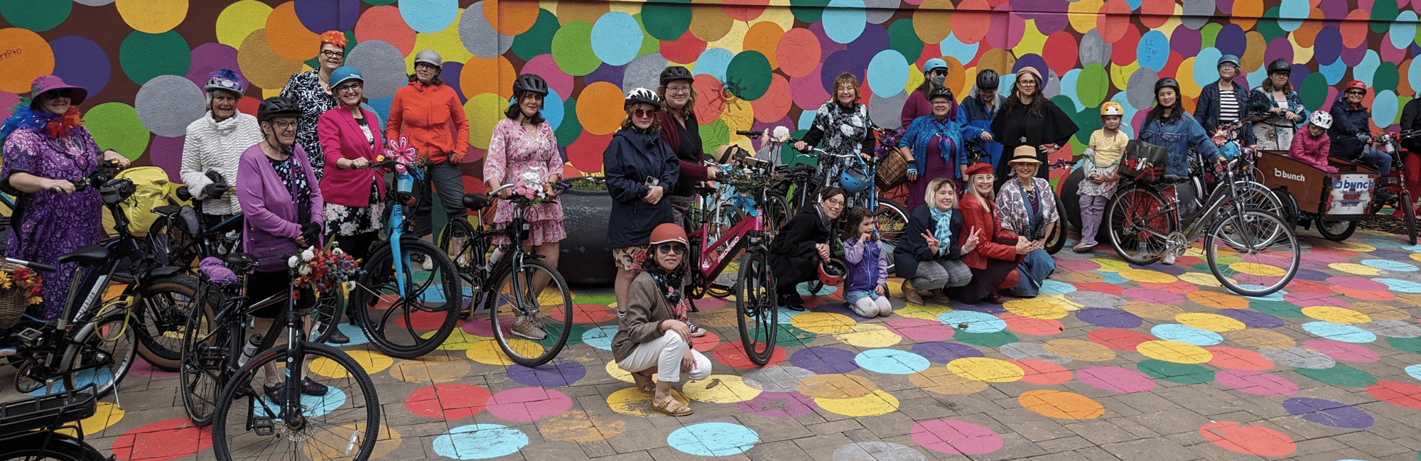 Many women, dressed up fancy, with some kids posing in front of an outdoor building mural of colourful dots with their bicycles