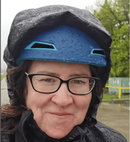 Sarah smiles on her bike in the rain, wearing a helmet and a rain cape, with rain on her face and glasses