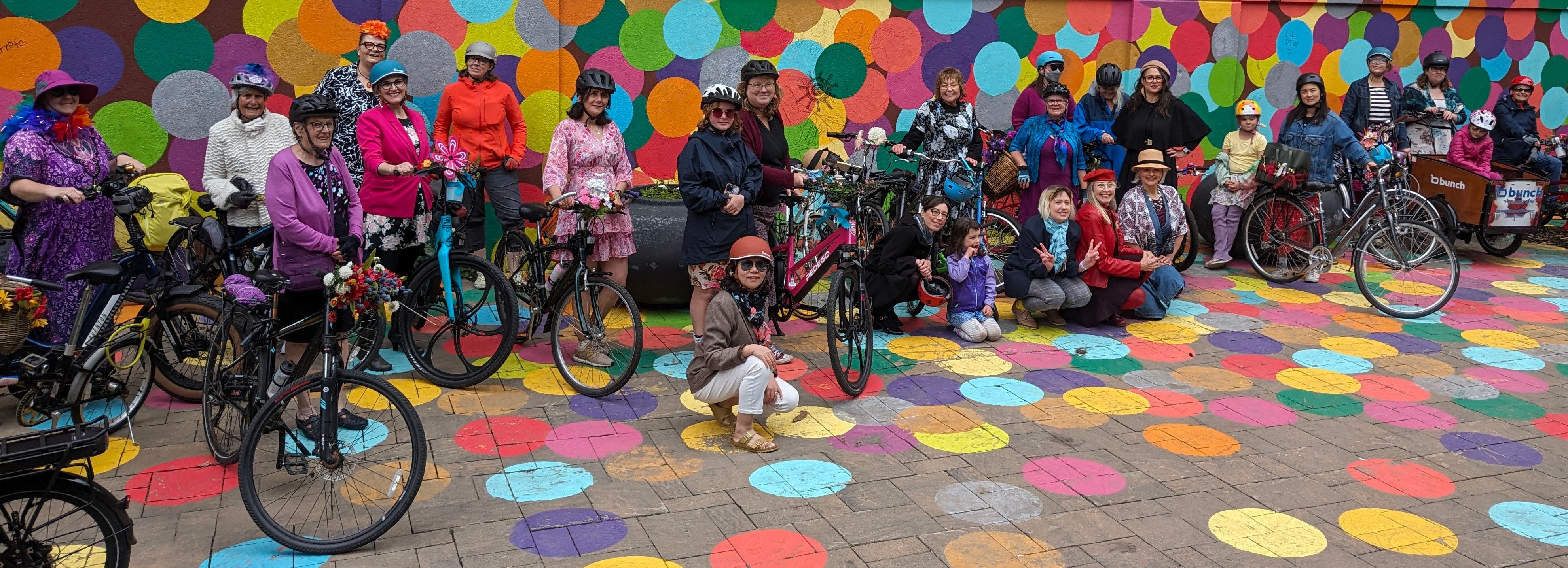 Many women, dressed up fancy, with some kids posing in front of an outdoor building mural of colourful dots with their bicycles