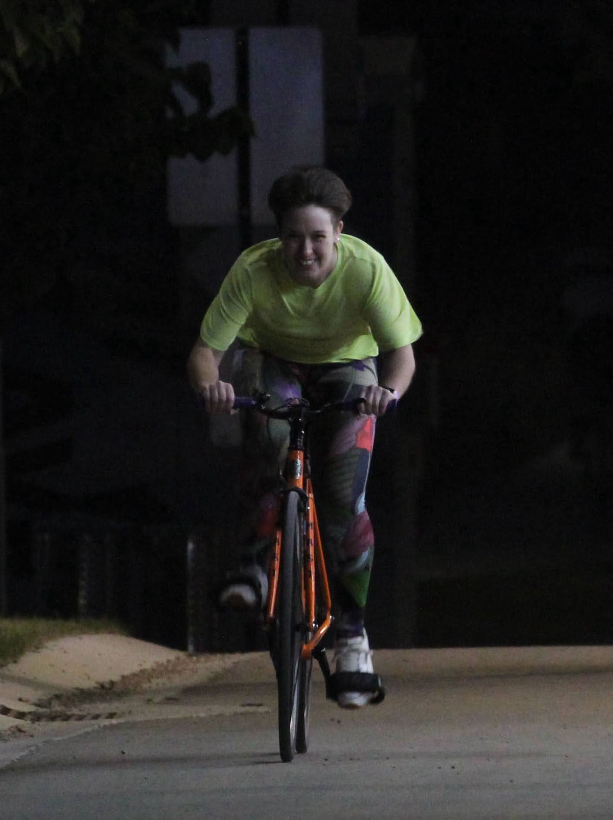 A person wearing a bright green t-shirt and multicoloured tights races their bright orange bike at night time