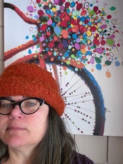 A photo of a woman wearing a red knit toque and black glasses in front of a poster on the wall of the front side profile of a red bicycle with a bike basket that is an explosion of colourful dots