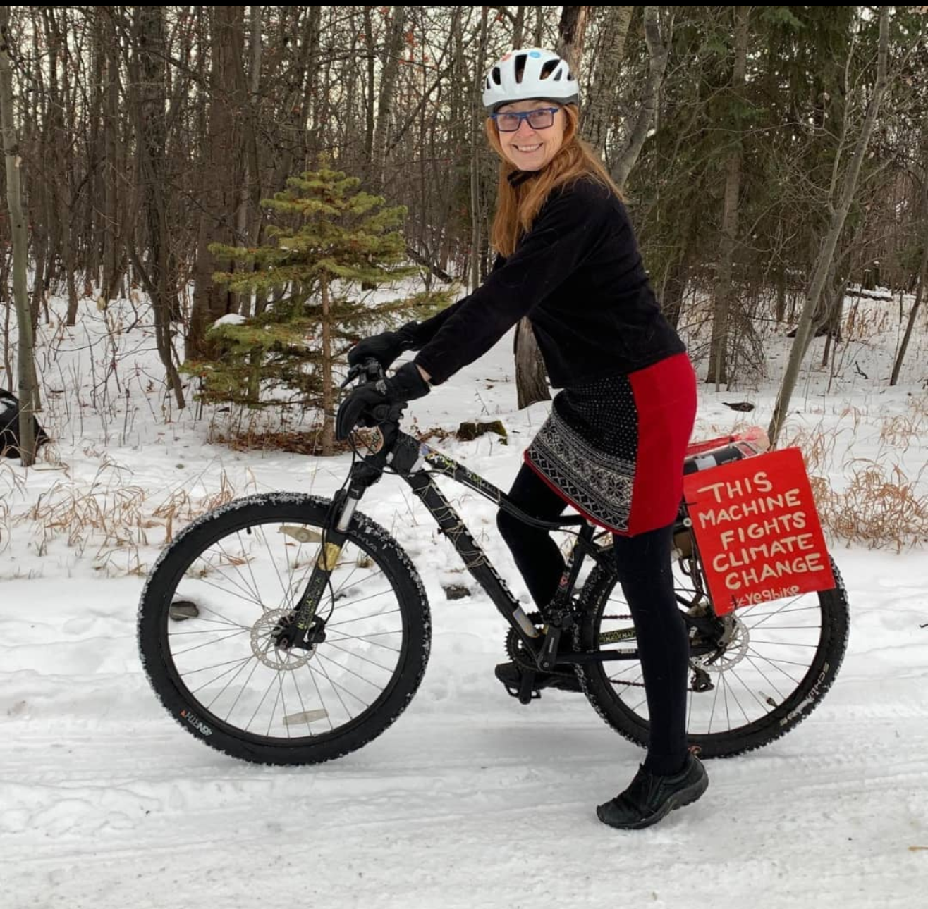 Woman with a white helmet on a bike in the winter wearing a JJWool skirt. There is a sign affixed on the rear of the bike that reads: This machine fights climate change