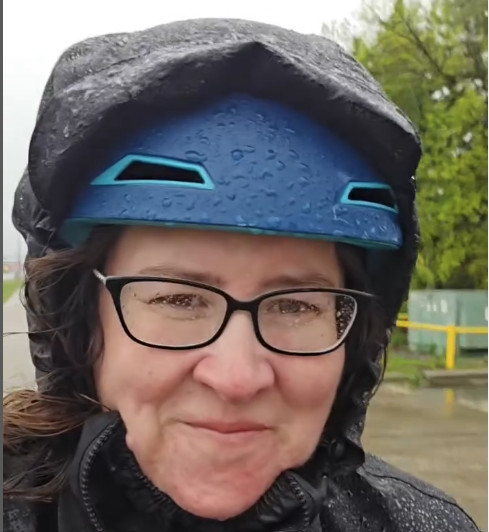 Sarah smiles on her bike in the rain, wearing a helmet and a rain cape, with rain on her face and glasses